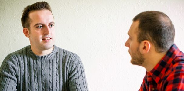 deux hommes discutent. L'un a un pull gris, l'autre une chemise à carreaux.